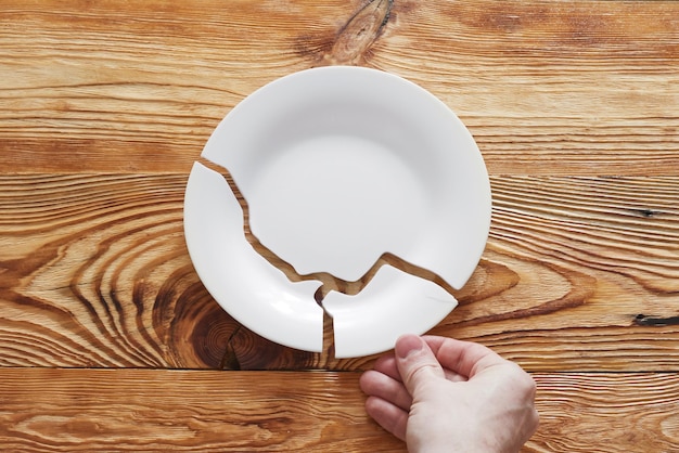 Top view on broken white plate and human hand holding one piece on wooden table | Premium Photo